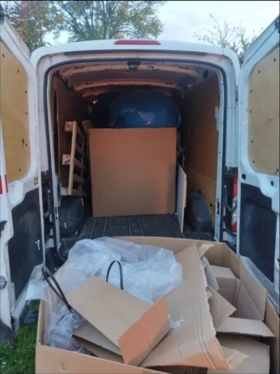 Wnętrze białego busa wypełnione kartonami, paletą i workiem. Widok z tyłu pojazdu z otwartymi drzwiami. Częściowo załadowany transport.