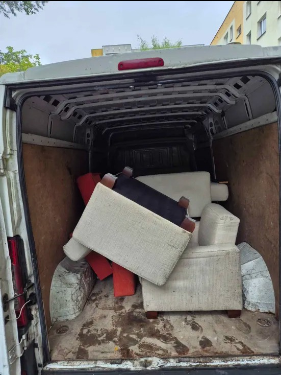 Wnętrze białego busa wypełnione różnokolorowymi pufami i fotelami z drewnianymi nóżkami. Widoczne ślady na podłodze i elementy karoserii pojazdu.