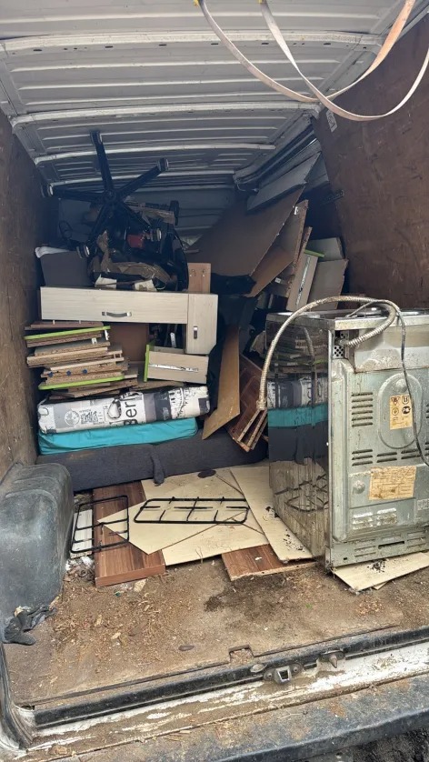 Wnętrze brudnego busa wypełnionego meblami, kartonami i sprzętem AGD, przygotowanego do transportu. Widoczny fragment kuchenki i biurowe krzesło.