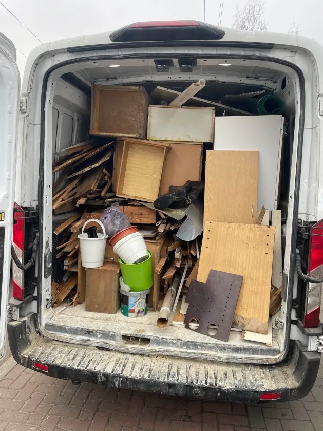 Wnętrze brudnego busa wypełnione gruzem, drewnem i odpadami po remoncie. Widoczne otwarte tylne drzwi i fragment chodnika.