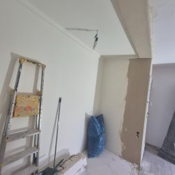 Remont pokoju: drabina, szpachlowana ściana, worek na gruz. Widoczne narzędzia i przygotowania do malowania. Fragment podłogi z parkietem.