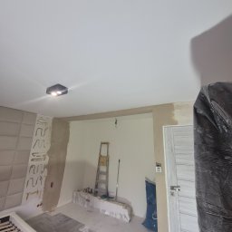 Pokój w trakcie remontu: częściowo położona podłoga, ściany przygotowane do malowania, widoczna drabina i zabezpieczone drzwi folią. Fragment łóżka w kadrze.