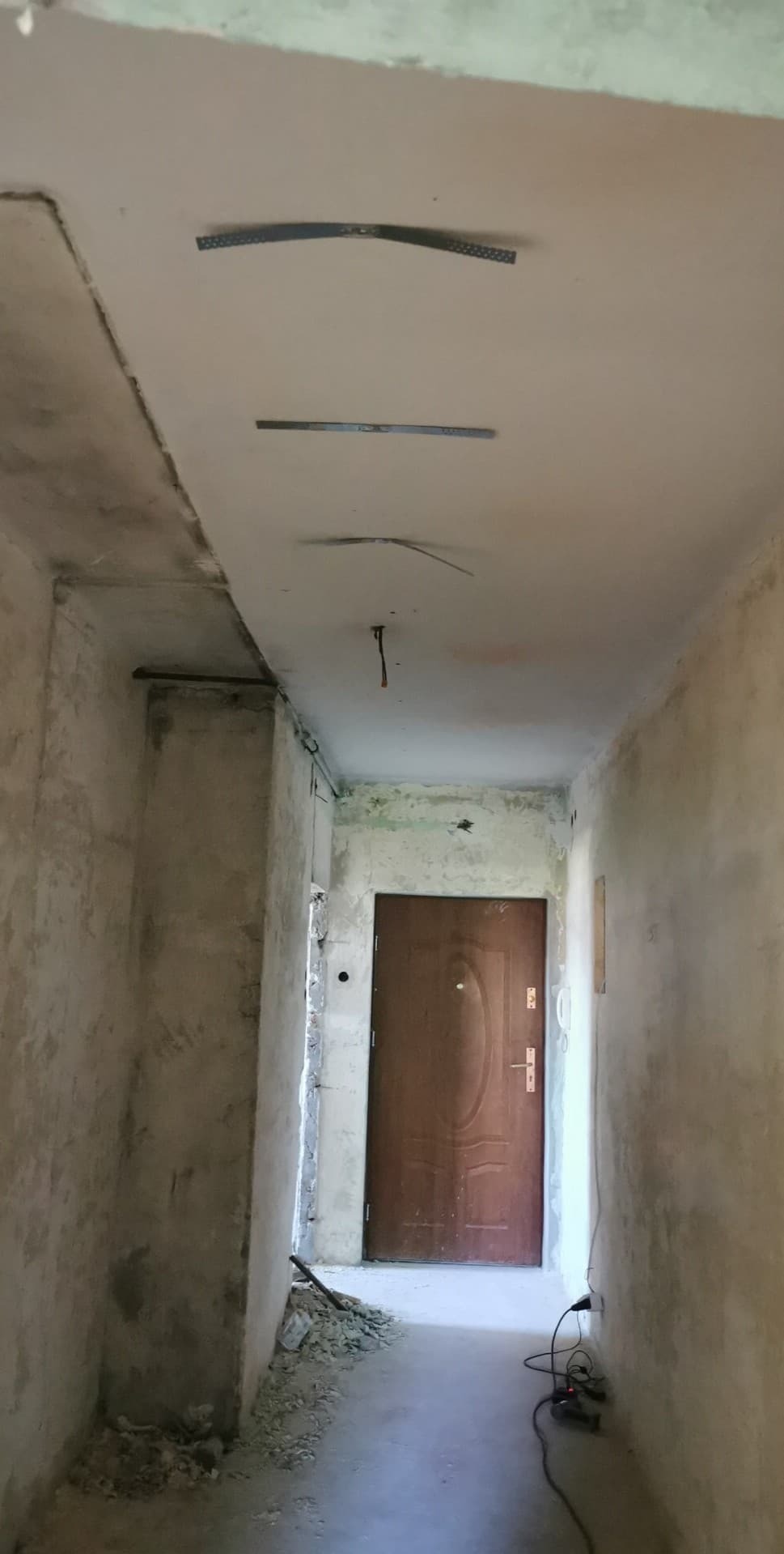 Korytarz w trakcie remontu: surowe ściany, widoczne elementy konstrukcyjne sufitu podwieszanego, przewody elektryczne i narzędzia na podłodze. Drzwi w tle.