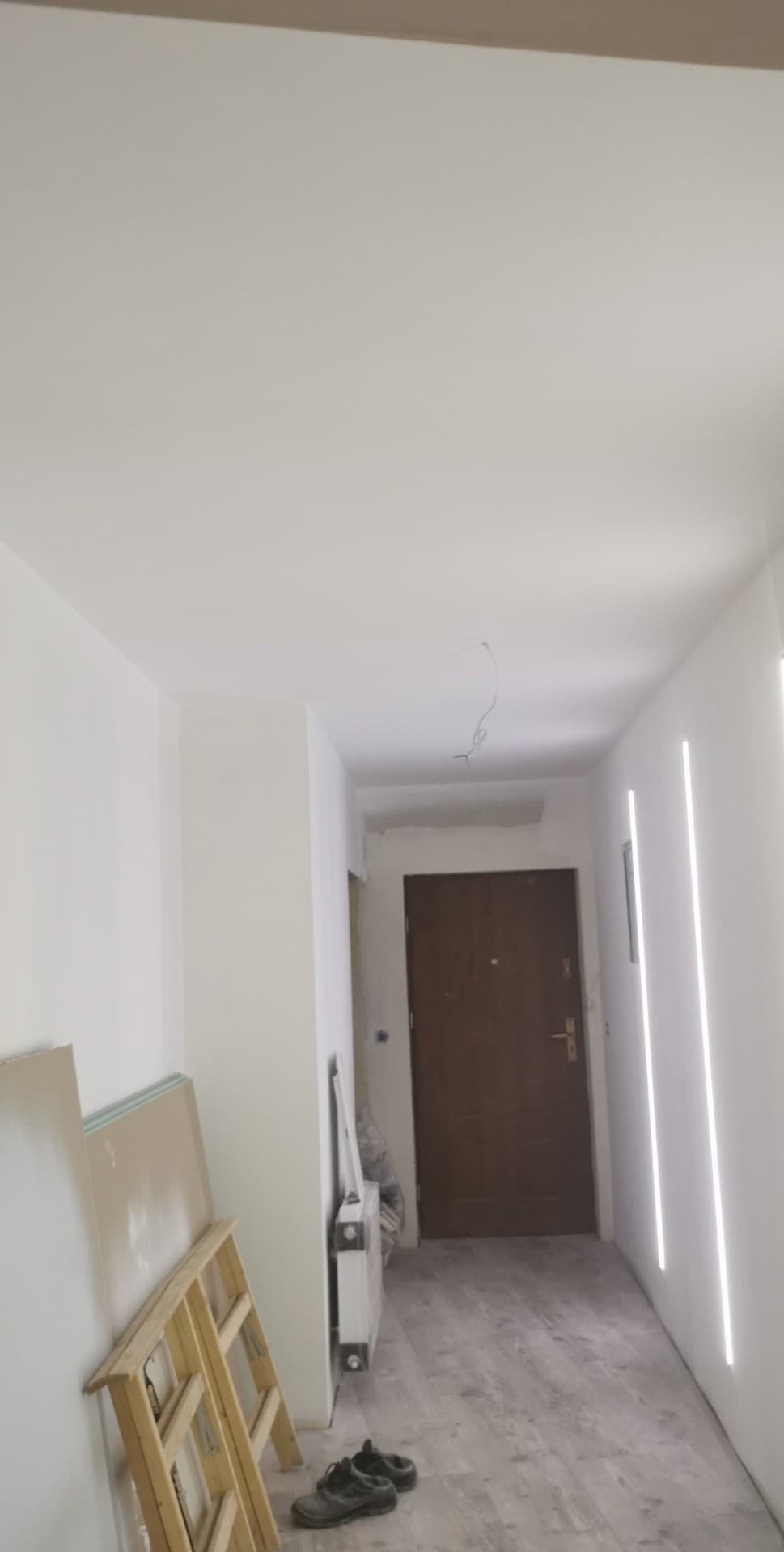 Białe wnętrze korytarza po remoncie z panelami LED na ścianie. Widoczne płyty karton-gips oparte o ścianę i drabina malarska. Podłoga wyłożona panelami.