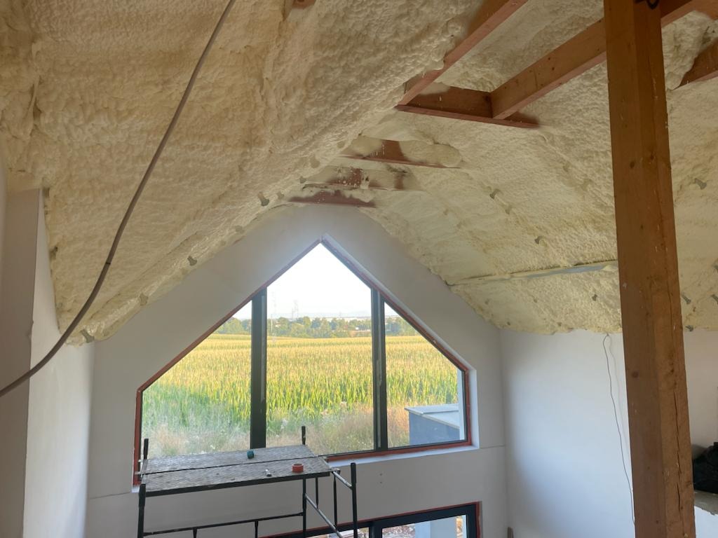 Poddasze z oknem i widokiem na pole, ocieplone pianą PUR. Widoczne drewniane elementy konstrukcyjne i rusztowanie. Świeżo ocieplone poddasze w trakcie prac wykończeniowych.