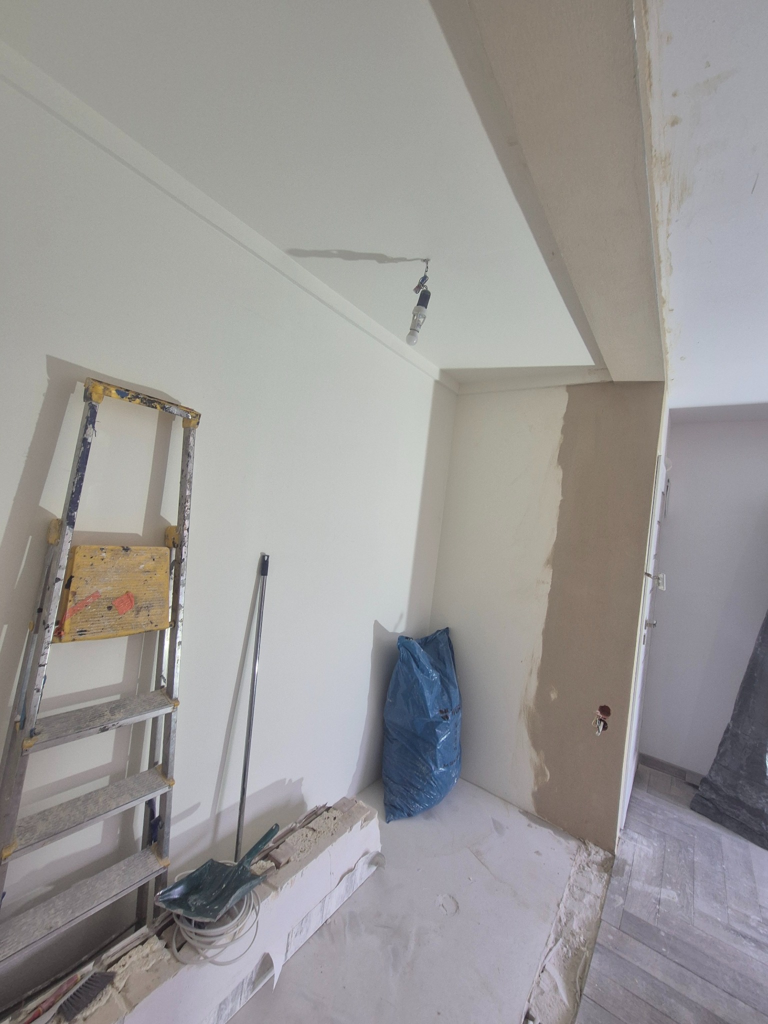 Remont pokoju: drabina, szpachlowana ściana, worek na gruz. Widoczne narzędzia i przygotowania do malowania. Fragment podłogi z parkietem.