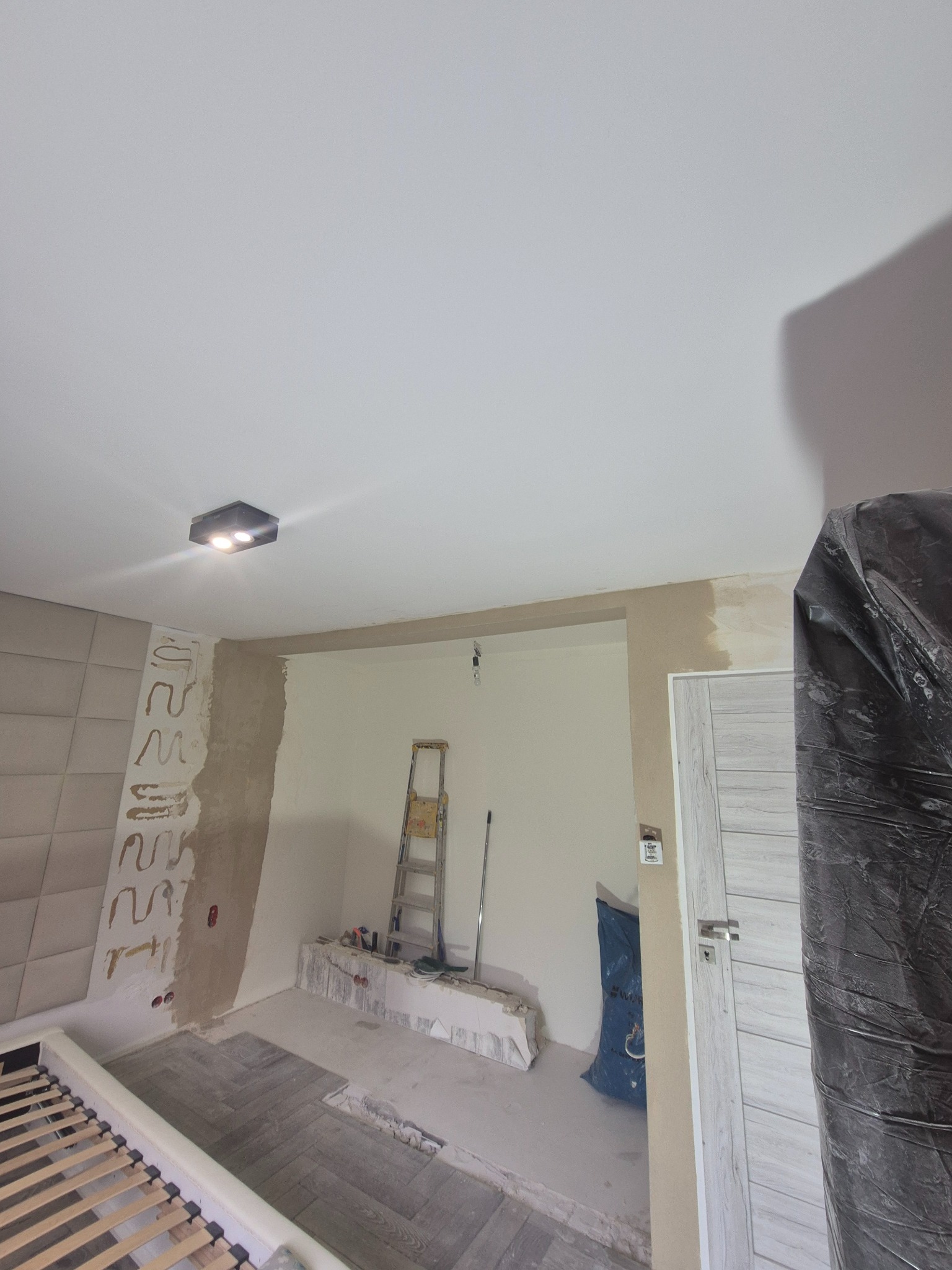 Pokój w trakcie remontu: częściowo położona podłoga, ściany przygotowane do malowania, widoczna drabina i zabezpieczone drzwi folią. Fragment łóżka w kadrze.