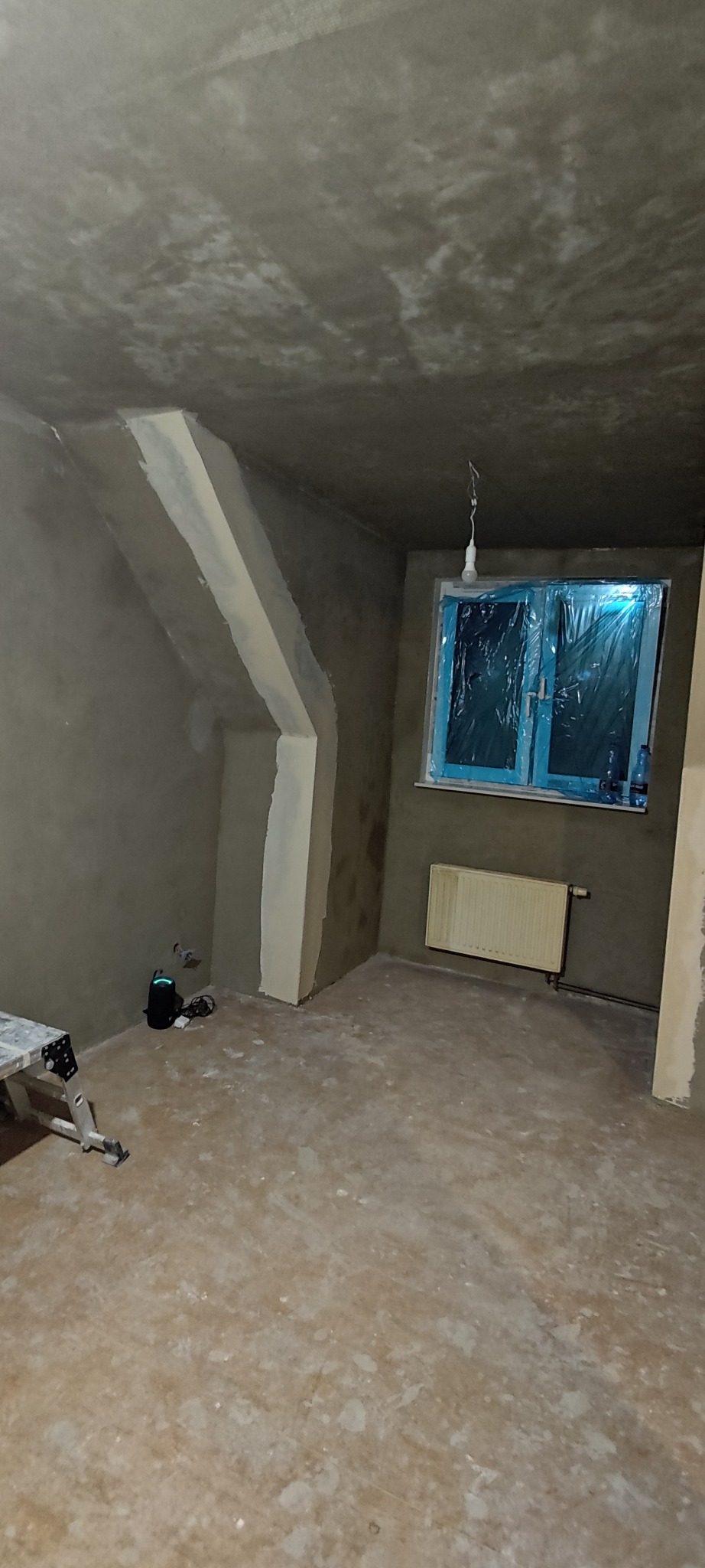 Pokój w trakcie prac wykończeniowych: ściany po gruntowaniu, zabezpieczone okno folią, widoczny grzejnik i drabina malarska. Przygotowanie do malowania.