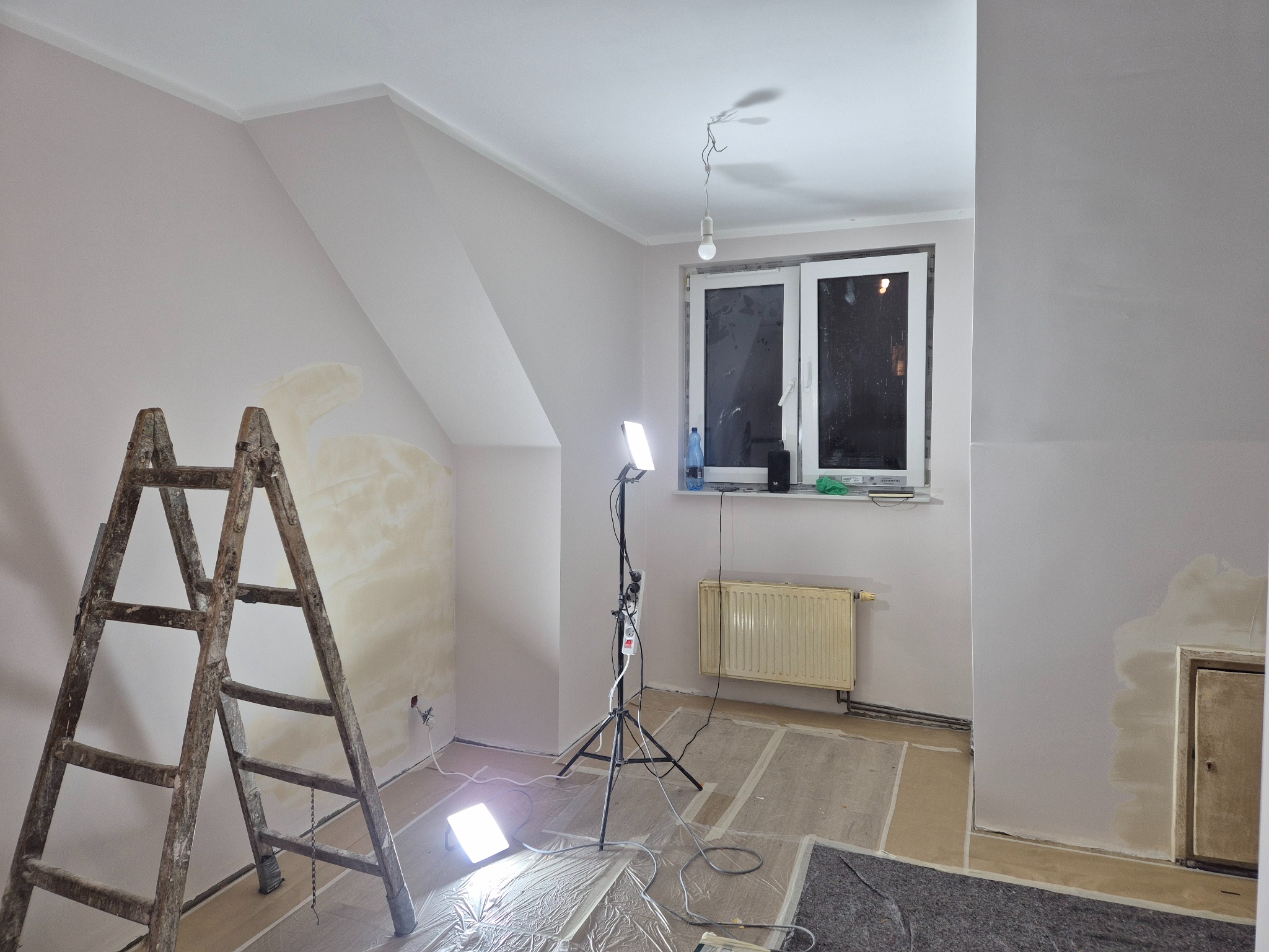 Pokój w trakcie prac wykończeniowych: ściany częściowo pomalowane na jasny kolor, drabina, lampa na statywie, zabezpieczona podłoga. Widoczne niedoskonałości i detale.