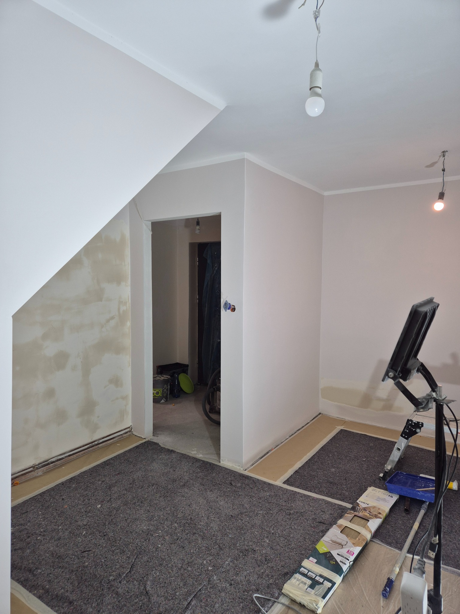 Wnętrze w trakcie malowania: zabezpieczona podłoga, ściany częściowo pomalowane na jasny odcień, widoczny sprzęt malarski i oświetlenie robocze.