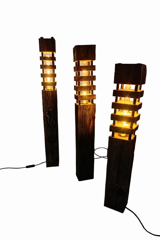 Trzy drewniane lampy podłogowe z ciepłym światłem, każda z unikalnym wzorem słojów i rozmieszczeniem elementów oświetleniowych, na białym tle.