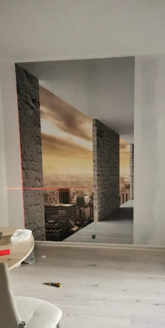 Fototapeta z motywem miejskiego krajobrazu, imitująca kamienne filary, zamontowana na ścianie. Widoczny laser poziomujący i narzędzia na podłodze.