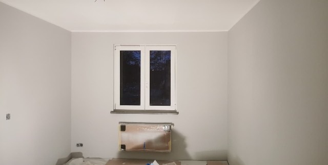 Jasnoszary pokój po malowaniu z białym oknem. Grzejnik zabezpieczony folią. Widoczne gniazdka elektryczne i fragment podłogi z folią ochronną.