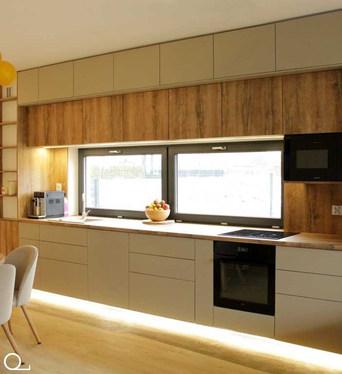 Nowoczesna kuchnia z drewnianymi szafkami górnymi i beżowymi dolnymi, oświetleniem LED pod szafkami i owocami w misce na blacie przy oknie.