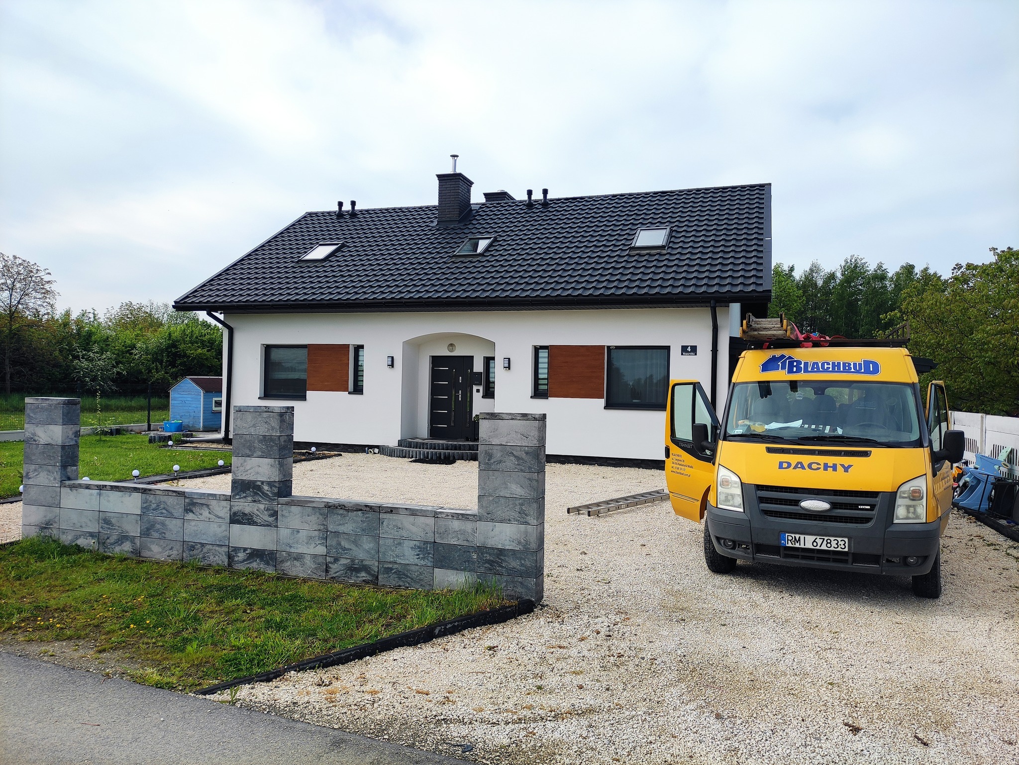 Nowoczesny dom jednorodzinny z czarnym dachem z dachówki ceramicznej i żółtym samochodem firmy dekarskiej z otwartymi drzwiami na żwirowym podjeździe.