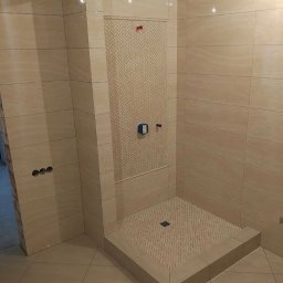 Aqua remont &secobau - Beżowa kabina prysznicowa z mozaiką w jodełkę na ścianie i podłodze, wykończona płytkami imitującymi kamień. Widoczne elementy armatury i odpływ liniowy.