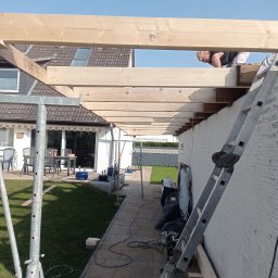 Aqua remont &secobau - Montaż drewnianej konstrukcji z belek na zewnątrz budynku. Widoczny pracownik na drabinie, elementy rusztowania i narzędzia. Prace w toku, jasne oświetlenie.