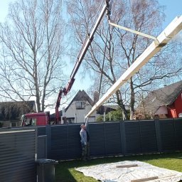 Aqua remont &secobau - Montaż drewnianej belki dźwigiem na tle szarego ogrodzenia i zielonego trawnika. Pracownik w jasnej kurtce nadzoruje proces. Widoczny fragment czerwonej ciężarówki.