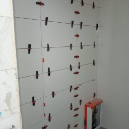 Aqua remont &secobau - Białe płytki ścienne w trakcie montażu, z użyciem klipsów poziomujących i krzyżyków dystansowych. Widoczna zabudowa rur w rogu pomieszczenia.