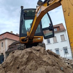 Aqua remont &secobau - Żółta koparka na stercie ziemi z gruzem, w tle budynki mieszkalne. Widoczna otwarta kabina operatora i ramię koparki. Prace ziemne w otoczeniu zabudowy.