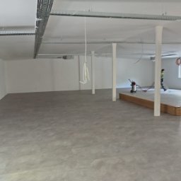 Aqua remont &secobau - Wykończone wnętrze biurowe z szarą podłogą, białymi ścianami i sufitem z widocznymi instalacjami. W tle pracownik z odkurzaczem przemysłowym.