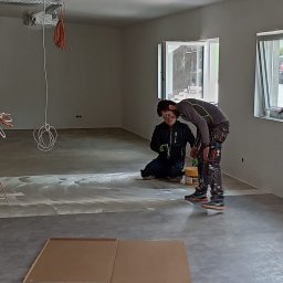 Aqua remont &secobau - Dwóch mężczyzn wyrównuje szpachelką podłogę w jasnym pomieszczeniu z dwoma oknami. Na podłodze leżą kartony ochronne. Widoczne kable pod sufitem.