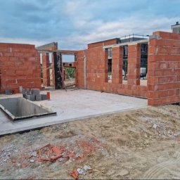 Aqua remont &secobau - Surowy stan budynku w trakcie budowy: czerwona cegła, betonowa podłoga, widoczne otwory okienne i drzwiowe, ziemia na zewnątrz. Niebo pochmurne.