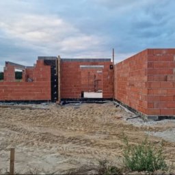 Aqua remont &secobau - Wznoszone mury domu z czerwonej cegły, widoczne otwory okienne i drzwiowe. Etap budowy w toku, z widocznym ociepleniem fundamentów i elementami konstrukcyjnymi.