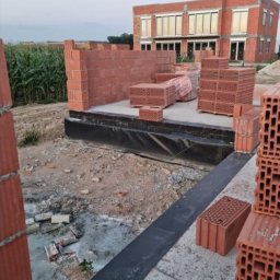 Aqua remont &secobau - Wznoszenie muru z czerwonej cegły na fundamencie z hydroizolacją. Częściowo postawiona ściana, palety cegieł i budynek w tle. Etap budowy domu jednorodzinnego.