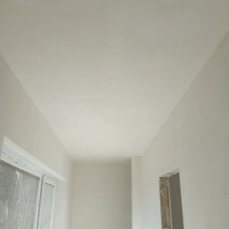 Aqua remont &secobau - Wnętrze po szpachlowaniu: gładkie, białe ściany i sufit, okno z folią ochronną, przygotowany otwór drzwiowy. Widok z dołu, podkreślający równość powierzchni.