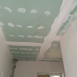 Aqua remont &secobau - Zielony sufit z płyt gipsowo-kartonowych, z widocznymi łączeniami i miejscami po wkrętach, przygotowany do szpachlowania. Widok z dołu, fragment okna w tle.
