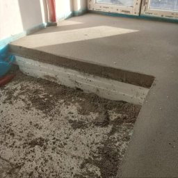 Aqua remont &secobau - Wyrównywanie podłoża w pomieszczeniu. Widoczna warstwa izolacji termicznej i wylewka betonowa. Fragment okna w tle. Prace przygotowawcze do położenia podłogi.