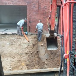 Aqua remont &secobau - Układanie rury w wykopie pod budynkiem z czerwonej cegły. Koparka w tle. Dwóch mężczyzn przy pracy. Etap prac ziemnych i instalacyjnych.
