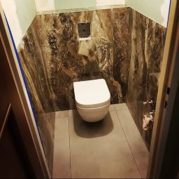 Aqua remont &secobau - Wykończona łazienka z podwieszaną toaletą, brązowe płytki imitujące marmur na ścianach, szare płytki na podłodze. Widok zza futryny drzwi.