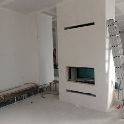 Aqua remont &secobau - Wnętrze w trakcie wykańczania: białe ściany, wnęka kominkowa z widoczną instalacją, drabina oparta o ścianę. Widoczne przewody i elementy konstrukcyjne.