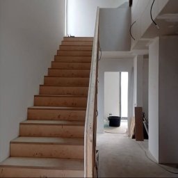 Aqua remont &secobau - Surowe, drewniane schody w jasnym wnętrzu w trakcie wykończenia. Widoczne przewody i wiadro, co sugeruje trwające prace. Minimalistyczny styl.