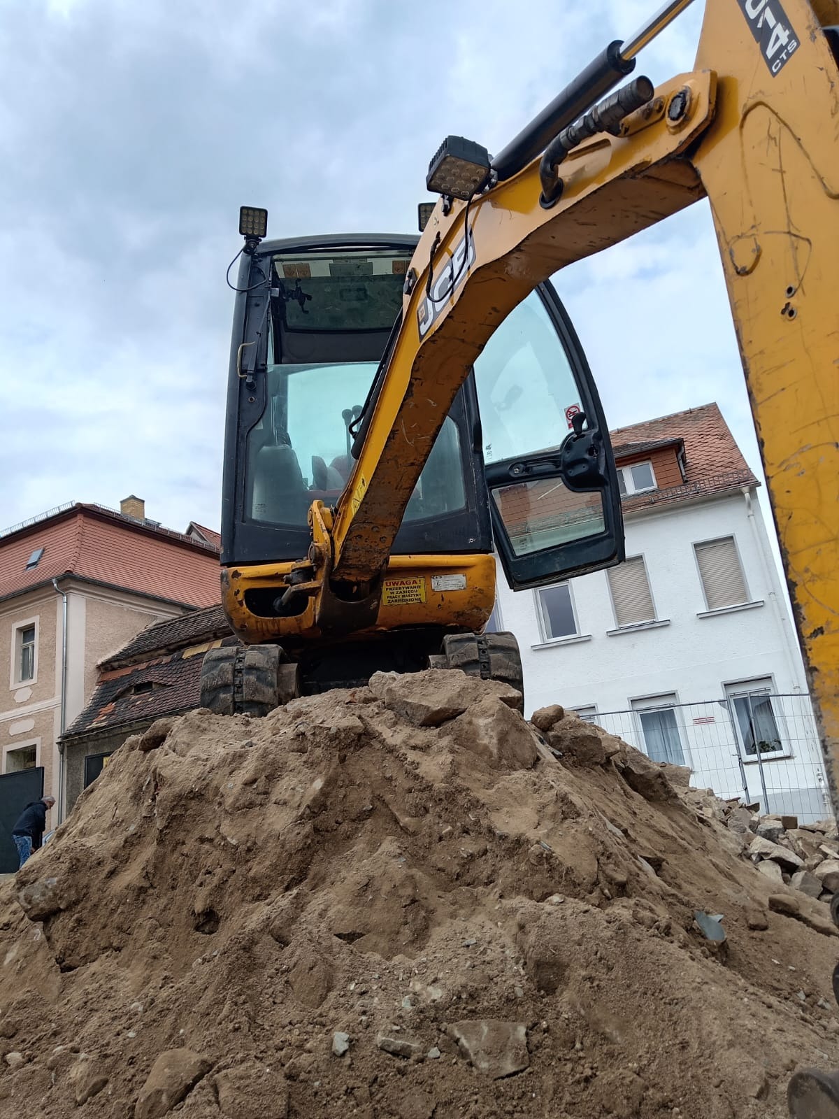 Żółta koparka na stercie ziemi z gruzem, w tle budynki mieszkalne. Widoczna otwarta kabina operatora i ramię koparki. Prace ziemne w otoczeniu zabudowy.