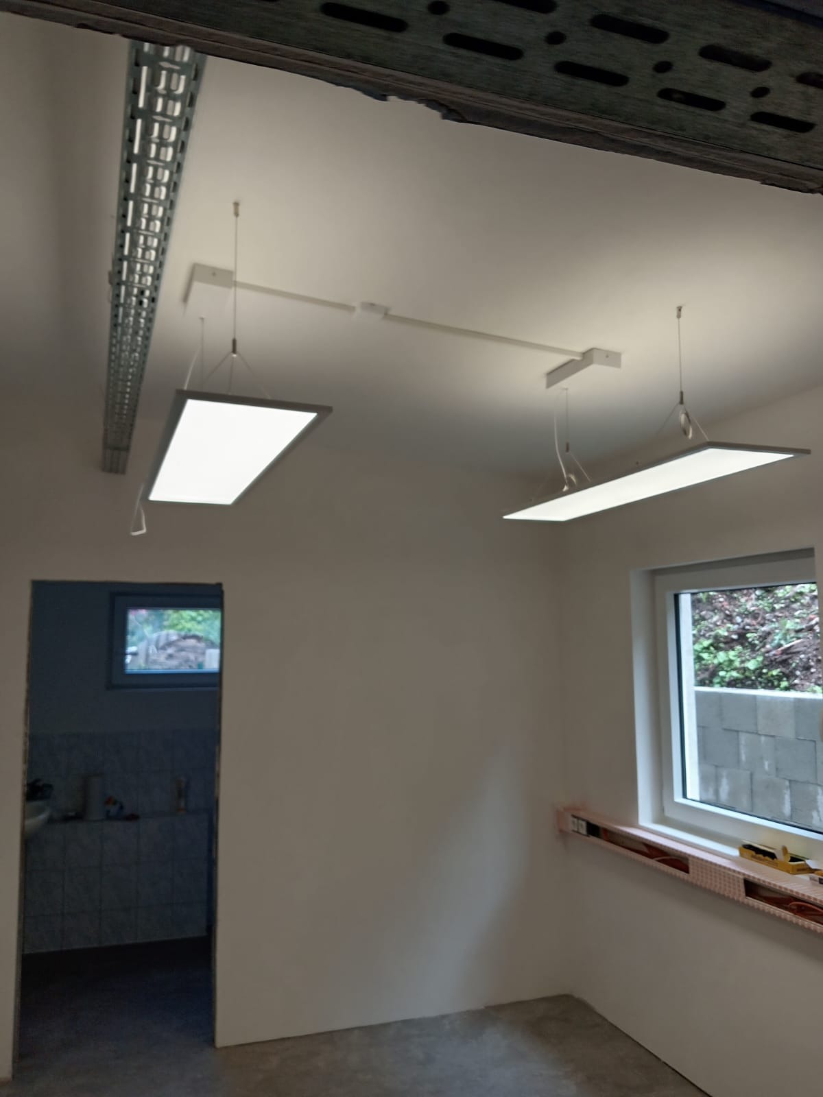 Dwa panele LED zawieszone na suficie w surowym wnętrzu. Widoczna instalacja elektryczna i okno z widokiem na zieleń. W tle wejście do pomieszczenia z wykończoną glazurą.