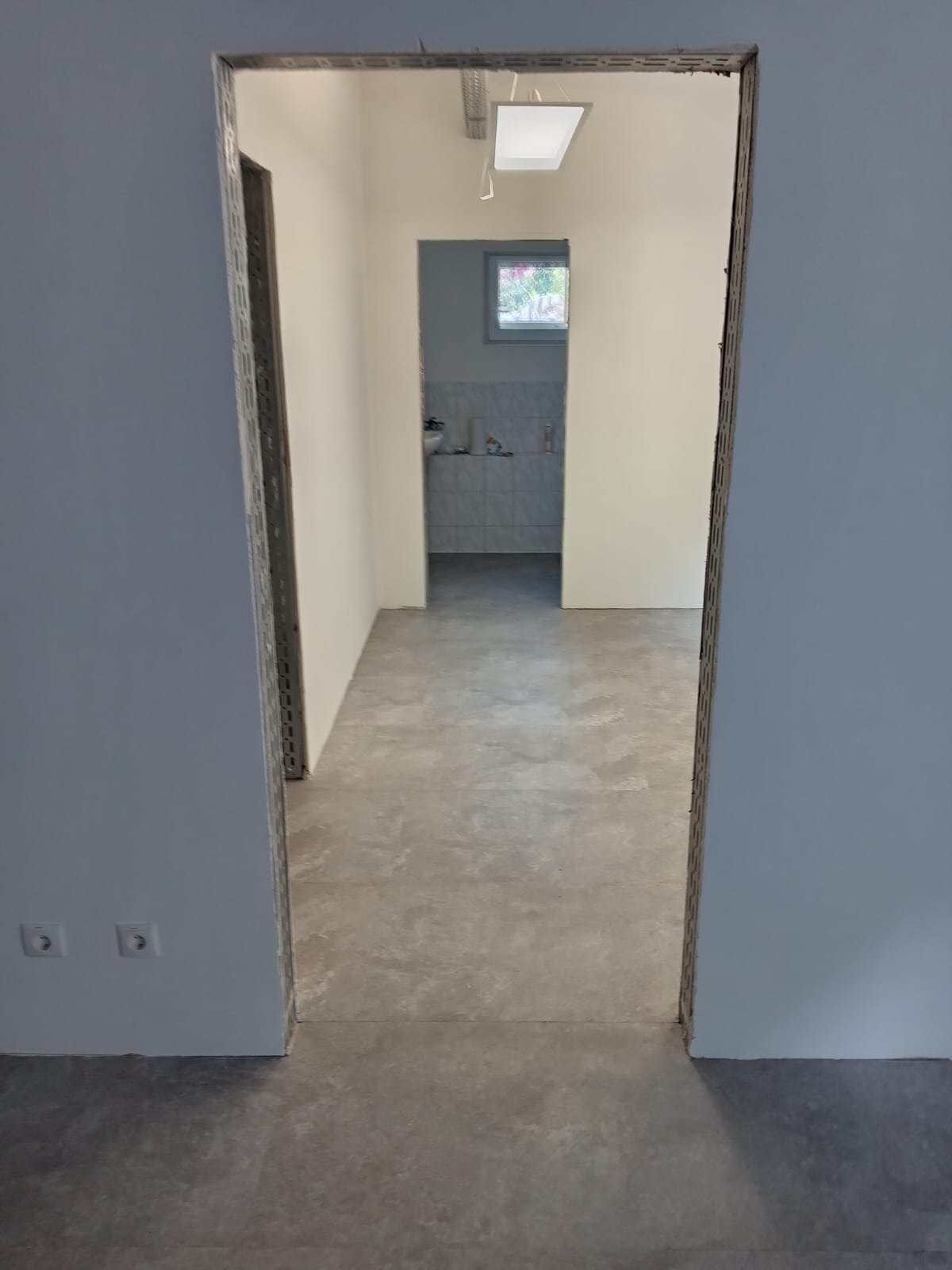 Wnętrze w trakcie wykańczania: szare płytki podłogowe, białe ściany, widok na łazienkę przez otwór drzwiowy z metalową ościeżnicą. Dwa gniazdka elektryczne po lewej.