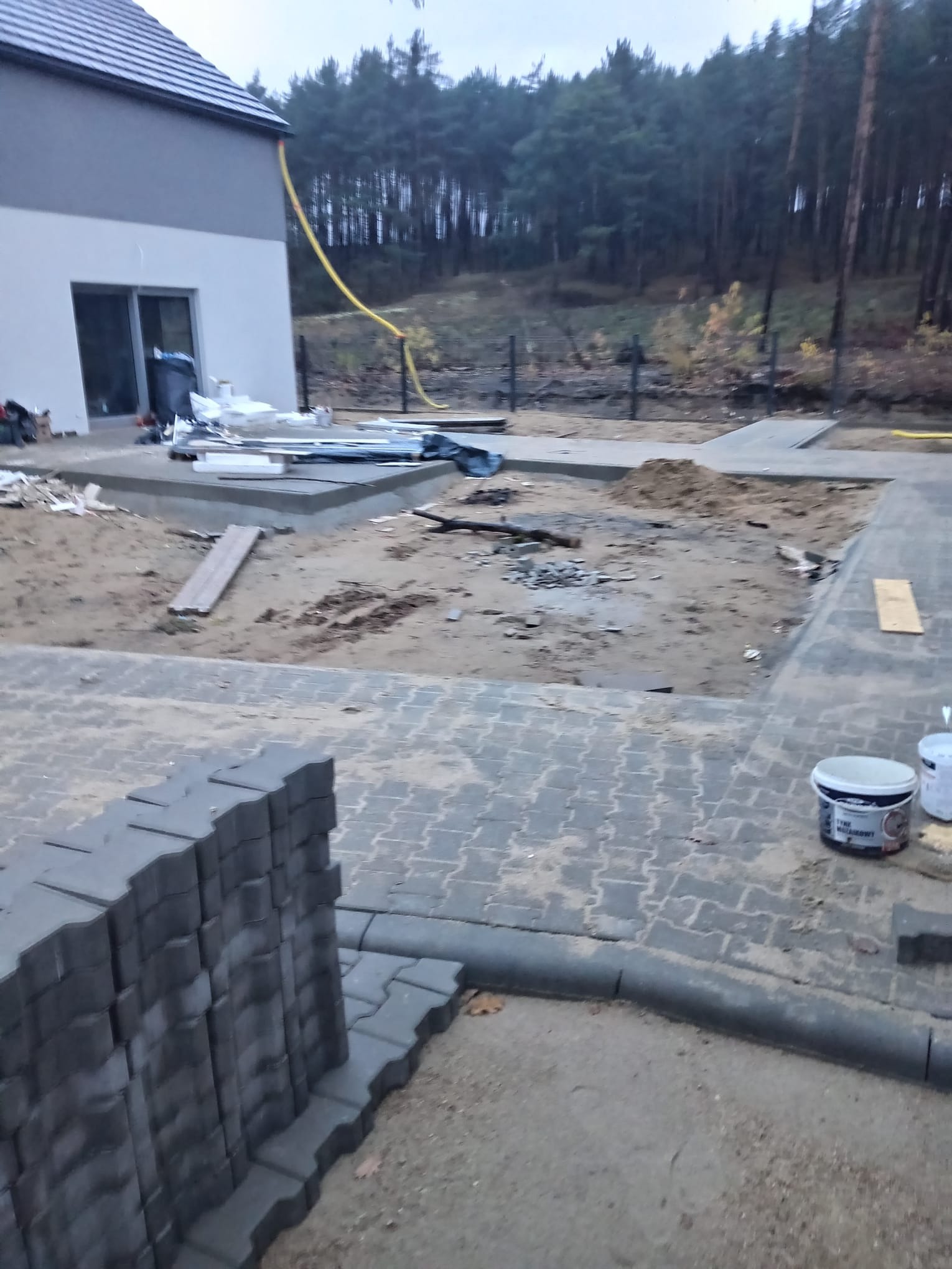 Układanie kostki brukowej na posesji domu jednorodzinnego. Widoczne ułożone fragmenty, stos kostki i narzędzia. W tle las i elewacja budynku.