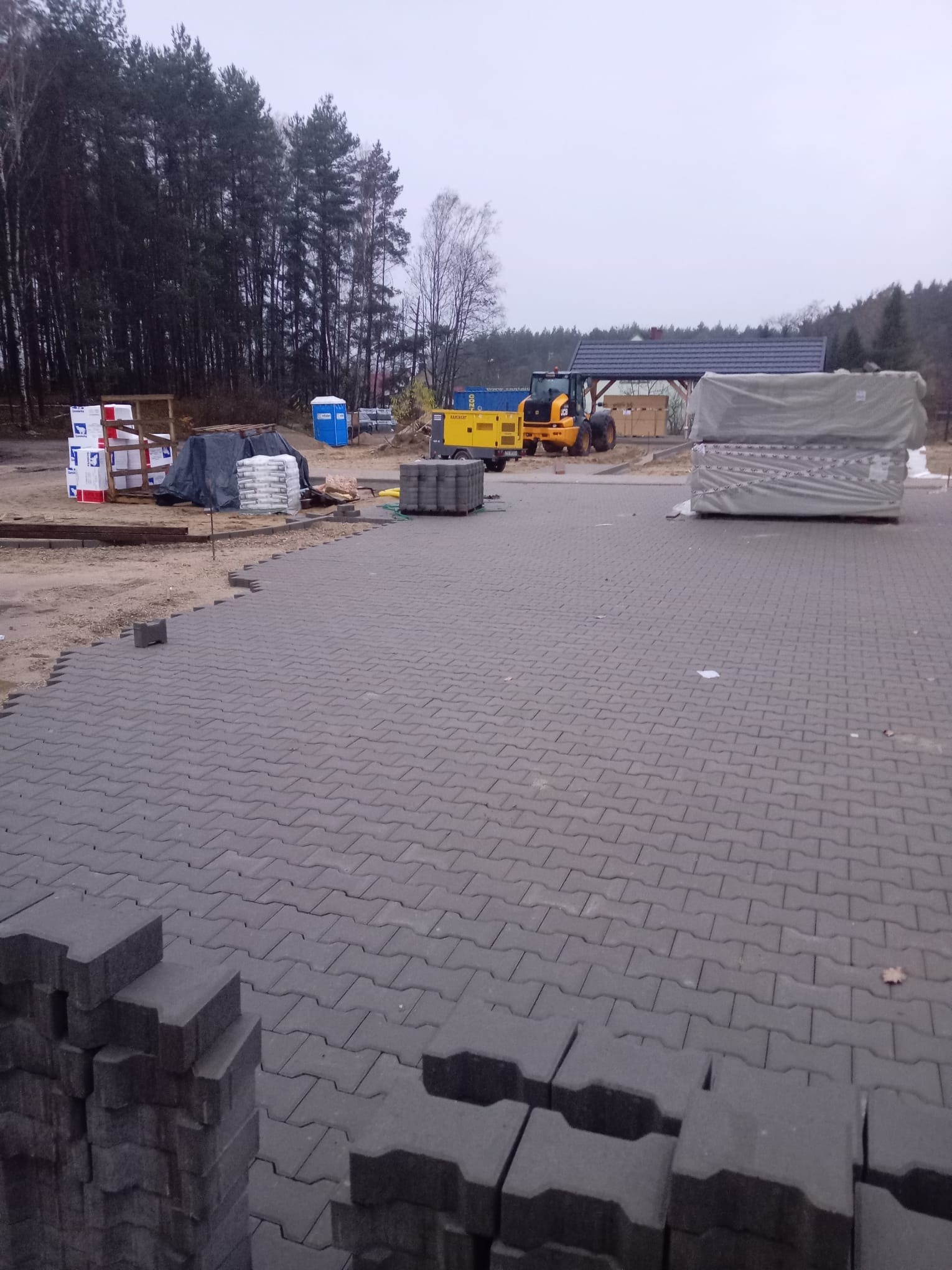 Układanie szarej kostki brukowej na posesji. W tle widoczny sprzęt budowlany i materiały. Częściowo ułożona nawierzchnia, stosy kostki czekają na montaż.