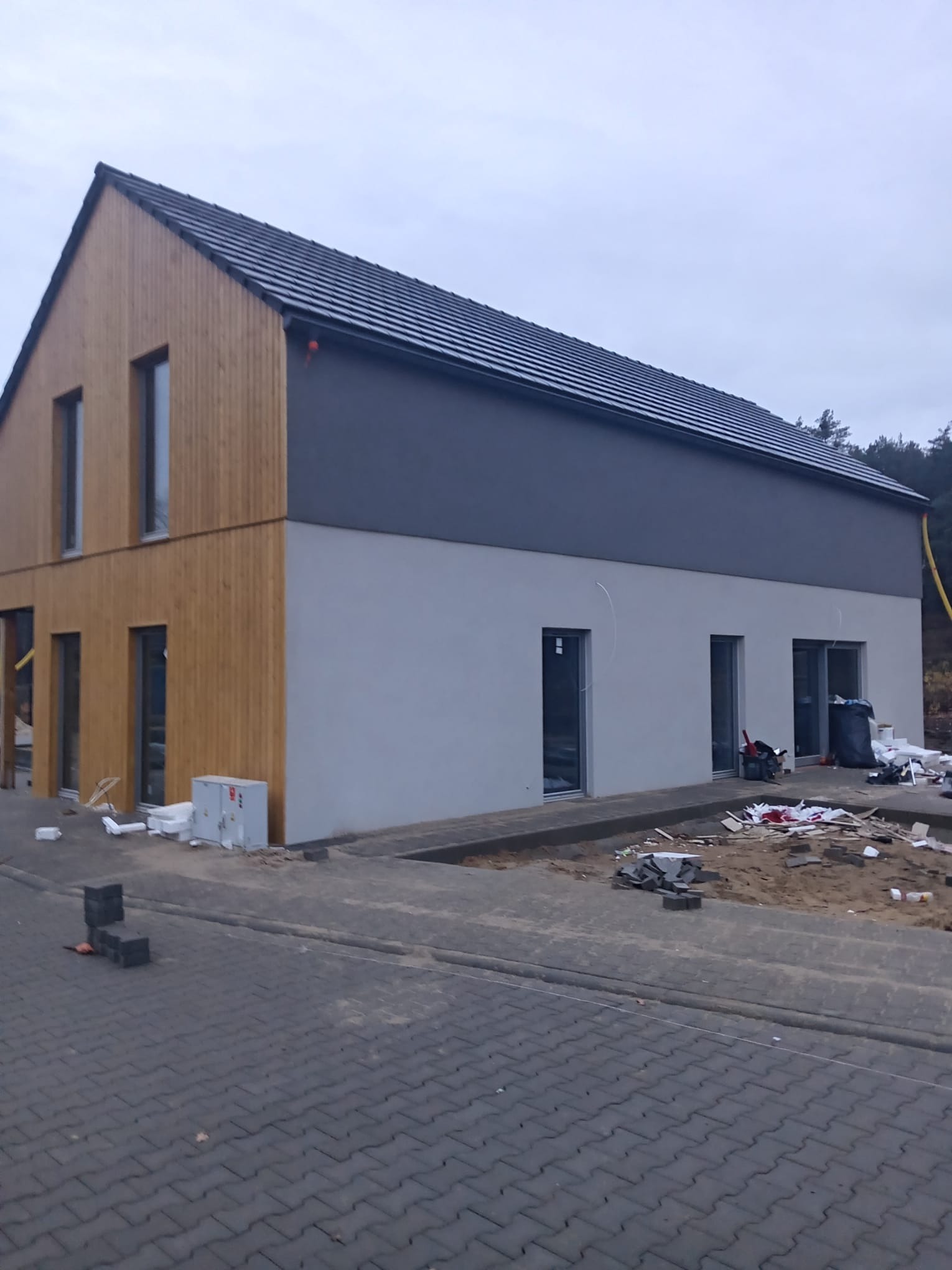 Dwukondygnacyjny dom z drewnianą elewacją i szarą górną częścią, w trakcie wykończenia. Widoczne materiały budowlane i kostka brukowa.