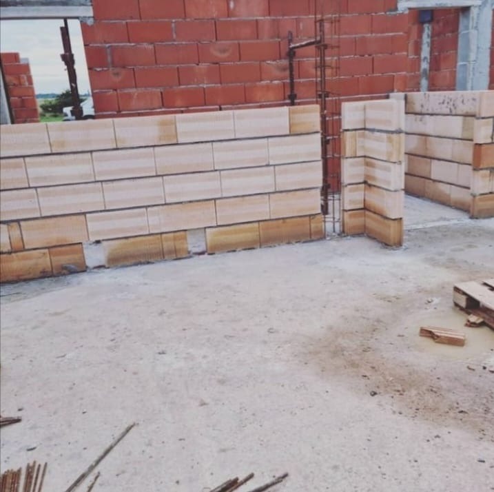 Wznoszenie ścian z bloczków ceramicznych, widoczne zbrojenie pionowe. Częściowo wymurowana ściana z otworami okiennymi. Budowa w toku, widoczne materiały budowlane.