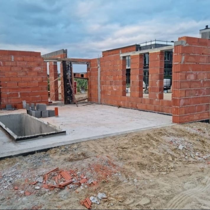 Surowy stan budynku w trakcie budowy: czerwona cegła, betonowa podłoga, widoczne otwory okienne i drzwiowe, ziemia na zewnątrz. Niebo pochmurne.