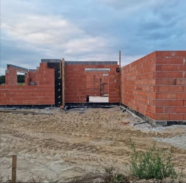 Wznoszone mury domu z czerwonej cegły, widoczne otwory okienne i drzwiowe. Etap budowy w toku, z widocznym ociepleniem fundamentów i elementami konstrukcyjnymi.