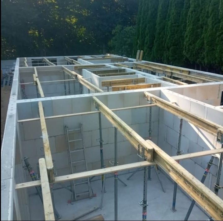 Wnętrze budynku w trakcie budowy z białych bloczków, wsparte drewnianymi belkami i metalowymi stemplami. Widoczna drabina i zieleń w tle.