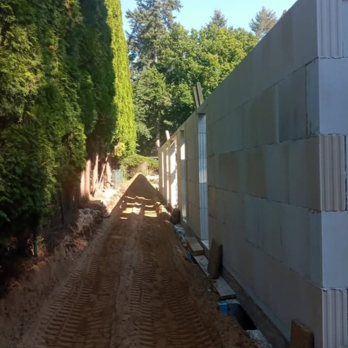 Ściana budynku w trakcie budowy z betonu komórkowego, widok z zewnątrz, ziemia z wykopu obok ściany, drzewa w tle, słoneczny dzień.