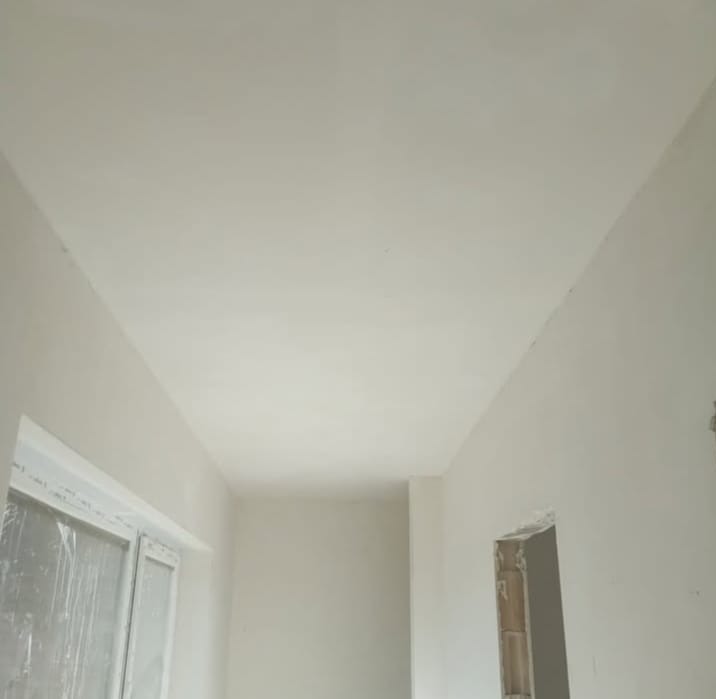Wnętrze po szpachlowaniu: gładkie, białe ściany i sufit, okno z folią ochronną, przygotowany otwór drzwiowy. Widok z dołu, podkreślający równość powierzchni.