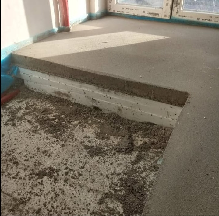 Wyrównywanie podłoża w pomieszczeniu. Widoczna warstwa izolacji termicznej i wylewka betonowa. Fragment okna w tle. Prace przygotowawcze do położenia podłogi.
