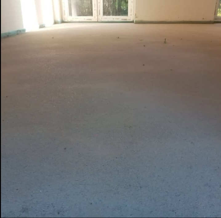 Wylana, gładka posadzka w jasnym pomieszczeniu z oknami balkonowymi. Widoczne zabezpieczenia ścian folią. Minimalistyczny kadr, skupiony na fakturze betonu.